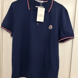 Marinblå Moncler pikétröja - Snygg marinblå pikétröja från Moncler med klassisk krage och korta ärmar. Tröjan har vita och röda ränder på krage och ärmslut samt broderad Moncler-logga på bröstet. Perfekt för en clean och sportig look.
