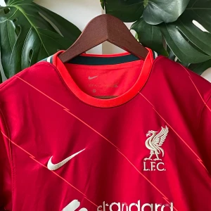 Liverpool FC matchtröja Nike - Liverpool FCs officiella matchtröja i rött med tunna diagonala linjer, broderad klubbmärke och Nike-logga på bröstet. Kortärmad modell med rund hals och orange detaljer vid kragen. Tillverkad i lätt och ventilerande material, perfekt för fotboll eller supporterliv.