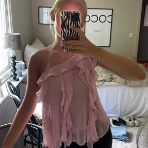 Ljusrosa volangig oneshoulder-top - Säljer en superfin ljusrosa oneshoulder-top med volanger. Toppen är ärmlös och har en luftig, flowy passform med flera lager volanger framtill. Använd endast en gång!