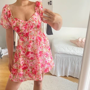 Blommig kortklänning med öppen rygg - Supersöt kortklänning med blommigt mönster i rosa, rött och vitt. Puffiga korta ärmar, söt v-ringning och öppen rygg med banddetalj. Perfekt för sommarens alla tillfällen, lätt och luftig känsla.