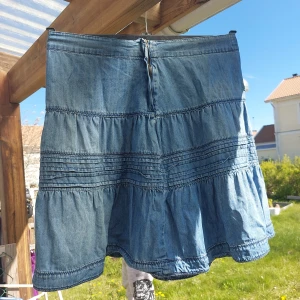 Blå volangkjol i jeans från Lindex - Supersöt blå jeanskjol från Lindex med volanger och dekorativa sömmar. Kjolen är i kort modell och tillverkad i 100% bomull, vilket gör den både mjuk och luftig. Perfekt för dig som gillar en avslappnad och trendig stil.