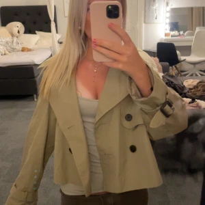 Beige croppad trenchcoat med knappar - Snygg croppad trenchcoat i beige med stora svarta knappar och bred krage. Jackan har bälte vid ärmsluten och en loose fit som ger en chill vibe. Perfekt för dig som vill ha en trendig och stilren look till våren eller hösten.