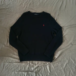 Mörkblå stickad tröja från Polo Ralph Lauren - Klassisk svart stickad tröja från Polo Ralph Lauren med rund halsringning och röd broderad logga på bröstet. Tröjan har ribbade muddar vid ärmslut och nederkant, perfekt för lager-på-lager under höst och vinter.