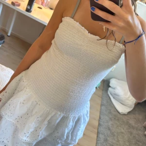 Vit sommarklänning från Gina Tricot - Supersöt vit kortklänning från Gina Tricot med smockad överdel och volangig kjol i broderad bomull. Axelbandslös modell som är perfekt för varma dagar. Klänningen har fina spetsdetaljer och en luftig känsla.