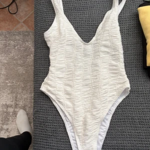 Vit baddräkt från Boohoo - Supersnygg vit baddräkt från Boohoo med rynkad struktur och djup v-ringning både fram och bak. Hög skärning vid benen ger en trendig look. Perfekt för stranden eller poolen, enkel att matcha med en sarong eller shorts. Har tvättat den därav är plomberingen borta men den var alldeles för liten på mig så väljer att sälja den. Helt oanvänd! 