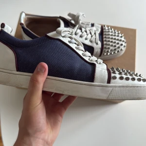 Christian Louboutin sneakers med nitar - Snygga sneakers från Christian Louboutin i marinblått tyg och vitt skinn, med ikoniska röda sulor och silverfärgade nitar på tån. Skorna har vita snören och röda detaljer längs kanterna. Perfekta för dig som vill sticka ut med en edgy och lyxig stil. Jag har använt dom en del det är därför under sulan är lite smutsigt och innersulan är ganska smutsig det är därför jag lägger ut dom för så billigt. Man får med låda dustbags och  extra nitar och dom är givetvis äkta