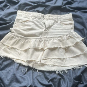 Vit volangkjol i jeans från Gina - Supersöt vit kort jeanskjol med volanger och råa kanter från Gina. Kjolen har klassiska jeansfickor fram och bak, dragkedja och knapp i midjan. Perfekt för dig som gillar en edgy men feminin stil.