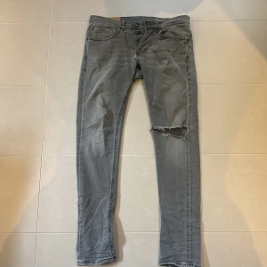 Dondup jeans  - Säljer ett par grå skinny jeans från Dondup i storlek 32. Jeansen har en snygg sliten look med ett stort hål på ena benet och klassiska fem fickor. Materialet är mjukt och stretchigt för en bekväm passform. Modellen är George 