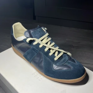 Maison Margiela blå sneakers mocka/läder - Maison Margiela sneakers i mörkblå mocka och läder med vita snören och beige sula. Klassisk low-top siluett med rund tå och platt gummisula. Kommer med originalbox och dustbags. Perfekt för dig som gillar stilrena och exklusiva sneakers.