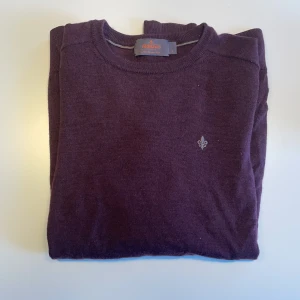 Vinröd merinoullströja från Morris - Vinröd stickad tröja från Morris i mjuk merinoull. Klassisk rund halsringning, diskret logga broderad på bröstet och ribbade muddar vid ärmslut och nederkant. Perfekt för lager-på-lager och snygg till jeans eller chinos.