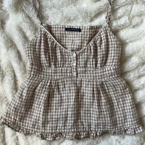 Rutig beige topp från Brandy Melville - Supersött rutigt linne i beige och vitt från Brandy Melville. Toppen har smala axelband, knappar framtill och volangkant nertill. Den är lätt utställd och har smock i ryggen för en bekväm passform. 