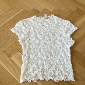 Vit spetsblus  - Supersöt vit blus i spets med korta ärmar och vågiga kanter. Toppens material är genombruten spets med ett unikt mönster som ger en romantisk vibe. Perfekt att styla med jeans eller kjol för en trendig look.