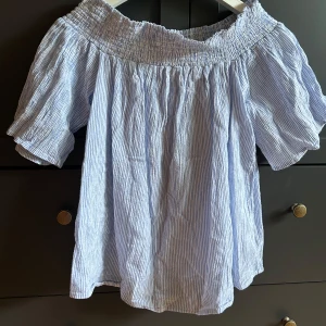 Randig offshoulder blus från Cavalini - Säljer en ljusblå och vit randig blus från Cavalini i storlek M. Blusen har offshoulder-modell med smock upptill och korta, luftiga ärmar. Perfekt för varma dagar med sin lätta bomullskänsla och avslappnade passform.