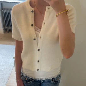 Krämvit kofta från Gina Tricot - Supersöt krämvit kofta från Gina Tricot med korta ärmar och rund hals. Koftan är stickad i mjukt material och har snygga knappar framtill. Perfekt att slänga över ett linne eller topp för en chill och stilren look.