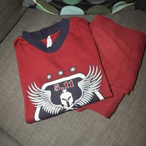 Röd 5.11 sweatshirt med tryck - Röd sweatshirt från 5.11 med coolt tryck framtill som visar vingar, hjälm och stjärnor. Tröjan har mörkblå ribbad krage och är gjord i mjuk bomull. Perfekt för dig som gillar sportig och avslappnad stil.