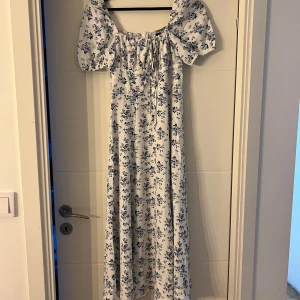 Vit långklänning med blå blommor - Superfin långklänning i vitt med blått blommönster och puffiga korta ärmar. Klänningen har snörning framtill och en romantisk vibe, perfekt för sommarens alla tillfällen. Sitter snyggt med lös passform och har en söt, feminin look. aldrig använd 