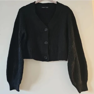 Svart stickad kofta från SHEIN - Svart ribbstickad kofta från SHEIN med croppad passform och stora knappar framtill. Koftan har v-ringning och långa ärmar med breda muddar. Perfekt för lager-på-lager och enkel att styla till olika looks.