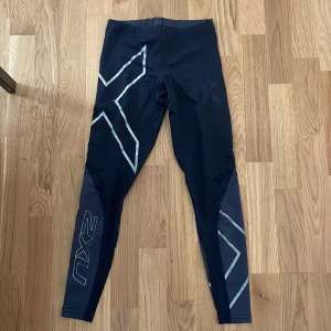 Svarta 2XU träningstights med reflex - Säljer ett par svarta träningstights från 2XU med silvriga reflexdetaljer längs benen. Tightsen har en tight passform och har låg och elastisk midja. 