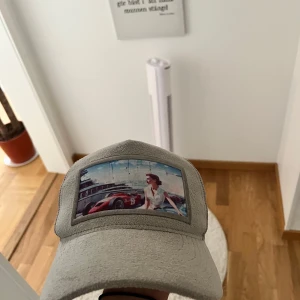 Grå meshkeps från Revenite med tryck - Snygg grå keps från Revenite med mesh på sidorna och baksidan för extra ventilation. Framsidan har ett coolt tryck med bil- och yachtmotiv. Perfekt för dig som vill sticka ut med en unik accessoar. Justerbar passform och böjd skärm.