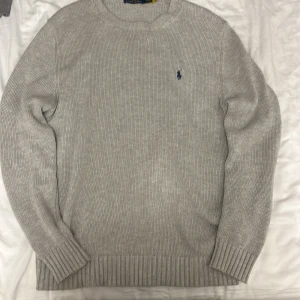 Grå stickad tröja från Polo Ralph Lauren - Snygg grå stickad tröja från Polo Ralph Lauren med klassisk rund hals och broderad marinblå logga på bröstet. Tröjan har ribbade muddar och är perfekt för lager-på-lager. Enkel att matcha och riktigt stilren.