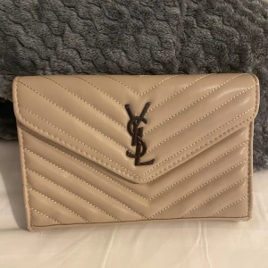 Beige YSL quiltad axelväska i skinn - Säljer en beige quiltad axelväska från YSL med klassiskt YSL-emblem i metall framtill. Väskan har kedjerem i mörk metall och är tillverkad i mjukt skinn. Snygg kuvertformad design med stilrena sömmar i sicksackmönster.