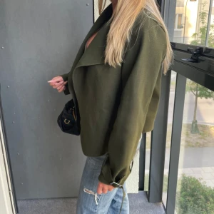 Kappa - Snygg olivgrön kappa med bred krage och bältdetaljer vid ärmsluten. Jackan har en loose fit och är perfekt att slänga över en t-shirt eller topp. Materialet känns mjukt och har en lätt struktur. Enkel att styla med jeans för en chill look.