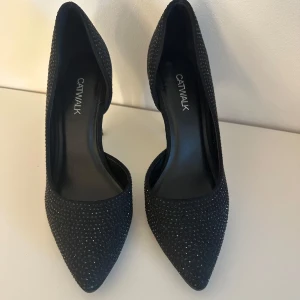 Svarta pumps med strass från Catwalk - Snygga svarta pumps från Catwalk med spetsig tå och täckta av små glittrande strasstenar. Köpa begagnat men säljs av fel storlek. Inget stort tecken på användning (bara på undersidan). Öppna på insidan så dom är lite luftiga. Köpta som storlek 38 men står 36 på undersidan. 