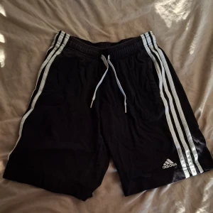 Svarta Adidas shorts med vita ränder - Svarta shorts från Adidas med klassiska vita ränder längs sidorna och vit snörning i midjan. Mjukt material och elastisk midja för skön passform. Perfekta för träning eller chill. Adidas-logga framtill. Barn shorts.