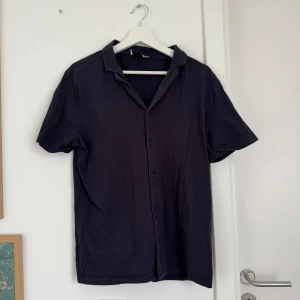 Svart kortärmad skjorta från Morris - Svart kortärmad skjorta från Morris i mjuk bomull. Klassisk krage, knäppning framtill och enkel, stilren design utan mönster. Perfekt för dig som gillar en clean och avslappnad look.