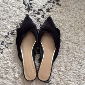 Svarta ballerinaskor/slip-ins i sammet från HM - Svarta ballerinaskor/slip-ins från HM i mjuk sammetsliknande material med spetsig tå och knutdetalj framtill. Platt sula och öppen häl. Endast provade inomhus. 