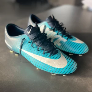 Nike Mercurial blå fotbollsskor - Snygga Nike Mercurial fotbollsskor i blått med svarta och vita detaljer. Skorna har randigt mönster, snörning och en stor vit swoosh på sidan. Perfekta för dig som vill sticka ut på planen. Lätta och smidiga med platt sula och dobbar för gräs.