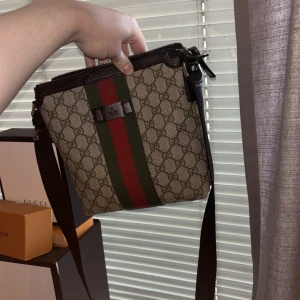 Gucci axelväska med GG-mönster - Säljer en Gucci axelväska i beige canvas med klassiskt GG-mönster och breda gröna och röda ränder framtill. Väskan har svart justerbar axelrem och detaljer i svart. Perfekt för dig som vill ha en ikonisk och trendig accessoar. Kvitto och box ingår 
