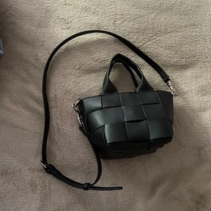 Svart flätad axelväska  - Säljer en svart axelväska i skinnimitation med flätad design. Väskan har både handtag och justerbar axelrem med silvriga detaljer. 