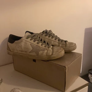 Beige sneakers från Golden Goose - Säljer ett par stilrena vita sneakers från Golden Goose med den ikoniska stjärnan på sidan. Skorna har svarta detaljer och snörning framtill. Perfekta för en avslappnad look. Tvättar såklsrt innan ja säljer för dome väldigt smutsiga på bilderna. Skriv om ni är intresserade