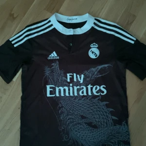 Real Madrid x Adidas fotbollströja - Snygg mörk fotbollströja från Adidas och Real Madrid med vit logga, vita ränder på axlarna och ett stort drakmotiv på framsidan. Tryck med Fly Emirates på bröstet och klassisk krage. Perfekt för dig som vill sticka ut på planen.
