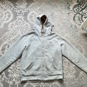 Grå hoodie från Burberry med dragkedja - Säljer en grå hoodie från Burberry med klassiskt rutigt foder i huvan. Tröjan har hel dragkedja, snörning i huvan och två stora fickor framtill. Perfekt för dig som gillar stilren design med en touch av lyx. Passar dem med storlek M men också L.