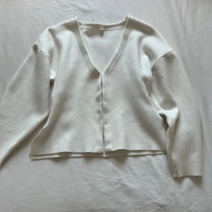 Vit kofta med v-ringning och slits - Minimalistisk vit kofta med v-ringad hals och diskreta hyskor som knäppning framtill. Långärmad modell i mjukt, stickat material med slitsar i sidorna för en chill och modern vibe. Perfekt att slänga över en topp eller klänning.