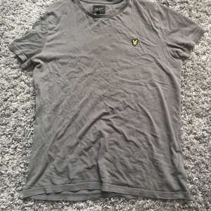 Grå t-shirt från Lyle & Scott - Snygg grå t-shirt från Lyle & Scott med klassisk rund hals och korta ärmar. T-shirten har den ikoniska gula loggan broderad på bröstet och är tillverkad i mjuk bomull. Perfekt basic-plagg med stilren look.