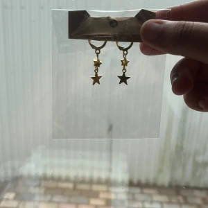 Guldiga örhängen med stjärnhängen - Snygga örhängen i guld med små hängande stjärnor. Perfekta för dig som vill ha en cool detalj till din look. Stjärnorna ger en trendig vibe och örhängena är lätta att matcha med andra smycken.