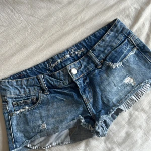 Blå jeansshorts från American Eagle - Snygga blå jeansshorts från American Eagle med slitna detaljer och fransig kant. Klassisk femficksmodell med låg midja och knappstängning. Insidan av benen har ett randigt tyg som syns när man viker upp kanten. Perfekta för sommaren!