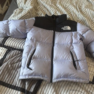 Ljuslila pufferjacka från The North Face - Säljer en snygg pufferjacka från The North Face i ljuslila med svarta axelpartier. Jackan har dragkedja framtill, två fickor med dragkedja och hög krage. Perfekt för kalla dagar och riktigt skön att bära. Logga på bröstet och ryggen.dne är en tjej model och en lite tajtare version💜