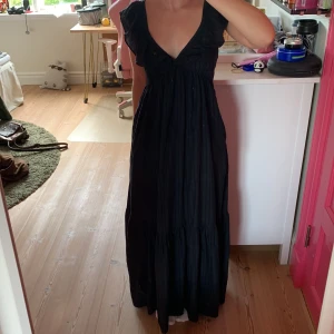 Svart långklänning med volang - Supersnygg svart långklänning med djup v-ringning och volangdetaljer vid axlarna. Klänningen har öppen rygg och är ärmlös, vilket ger en luftig och chic känsla. Perfekt för dig som vill ha en stilren och elegant look med lite extra edge.