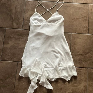 Vit kort klänning med volang från Zara - Supersöt vit kort klänning från Zara med tunna axelband som korsas i ryggen. Klänningen har volangdetalj nertill och en lätt satinliknande känsla. Perfekt för sommarens alla tillfällen när du vill känna dig extra fin. Finns någon form av fläck (se bild 3, vänster sida där axelbandet o klänningen är ihopsydda), syns dock knappt när den bärs. 