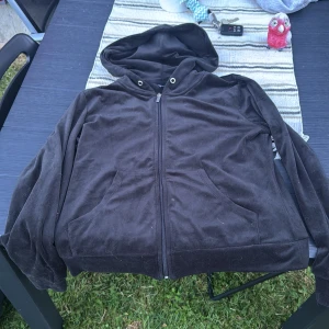 Svart hoodie med dragkedja  - Mjuk svart hoodiejacka i sammet med dragkedja framtill och två stora fickor. Jackan har huva med metallöljetter och långa ärmar. Perfekt för en chill och avslappnad stil.