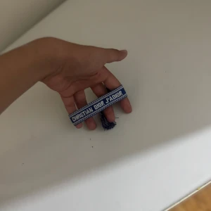 Blått armband Christian Dior J'Adior - Snyggt blått tygarmband från Christian Dior med texten 'J'Adior' broderad i vitt. Armbandet har justerbar knytning och tofsar i ändarna för en cool och avslappnad look. Perfekt accessoar för att lyfta din outfit med en touch av lyx.