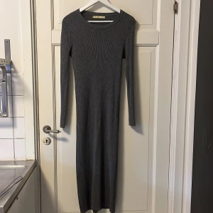 Grå ribbad långklänning  - Mörkgrå långklänning från Just Cute i ribbad stickad kvalitet. Klänningen har lång ärm, rund halsringning och en tight och figurnära passform som framhäver siluetten. Materialet är extremt mjukt och mysigt. Perfekt för dig som gillar en enkel men stilren look.