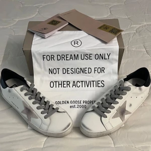 Golden Goose skor - Säljer ett par vita Golden Goose med en fin grå stjärna på sidan, svarta detaljer vid hälen och härliga gåta snören. Skorna har en klassisk låg sneakersmodell och är tillverkade av skinn med slitna detaljer för et mer avslappnat utseende. Perfekt för nuvarande sommar men även kommande höst och vinter. Ett till ett .Skorna är helt nya och säljs på grund av fel storlek. Nypris ligger på ca 5500kr. Vid några frågor är det bara att skriva 🤗. 