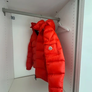 Röd Moncler pufferjacka med huva - Säljer en riktigt fet röd pufferjacka från Moncler med huva och dragkedja. Jackan har Moncler-logga på ärmen, stora fickor och coola detaljer. Perfekt för kalla dagar när du vill sticka ut och hålla värmen.