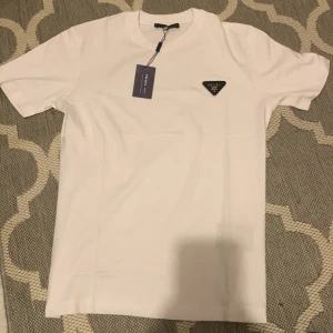 Vit t-shirt från Prada med logga - Vit t-shirt från Prada i klassisk modell med korta ärmar och rund hals. På bröstet sitter en svart, triangelformad Prada-logga i metall och tyg. T-shirten är gjord i mjuk bomull och har en clean, stilren look som passar till allt. Har aldrig haft på mig den, fick den idag, vill sälja den för den inte passar.