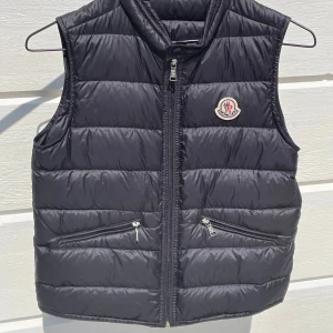 Svart dunväst från Moncler - Snygg svart dunväst från Moncler med klassisk logga på bröstet. Västen har quiltad design, två dragkedjefickor framtill och hög krage. Perfekt att slänga över en hoodie eller tröja för extra värme. Stängs med dragkedja framtill.